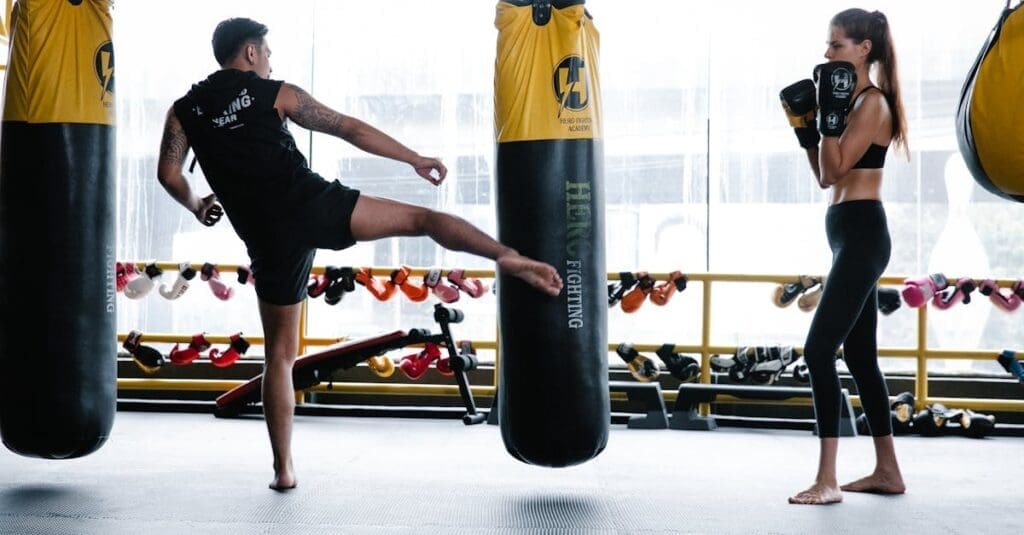 A man practices heavy bag kicks while a woman observes in boxing gloves, an essential drill from a beginner kick boxing guide.